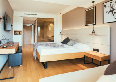 Blick in ein Zimmer im Hotel Kowald in Loipersdorf. Zu sehen ist ein Bett mit Holzrahmen, ein Beistelltisch aus Holz, Glaswände im Badezimmerbereich.