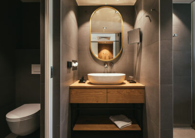 Blick in ein Badezimmer im Hotel Kowald in Loipersdorf. Zu sehen ist ein Waschtisch aus Holz, Steinfliesen an den Wänden, Spiegel, ein WC und ein Duschbereich.