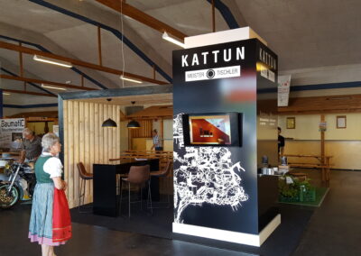 Kattun-Ausstellungsstand in der Martinihalle in Sankt Martin an der Raab. Offener Raum mit Tisch und Stühlen, Werbesäule mit Monitor.
